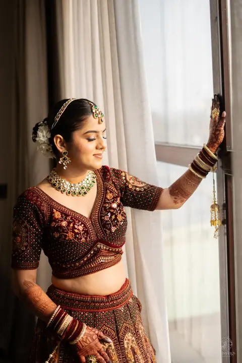 A bride in a maroon lehenga by a window